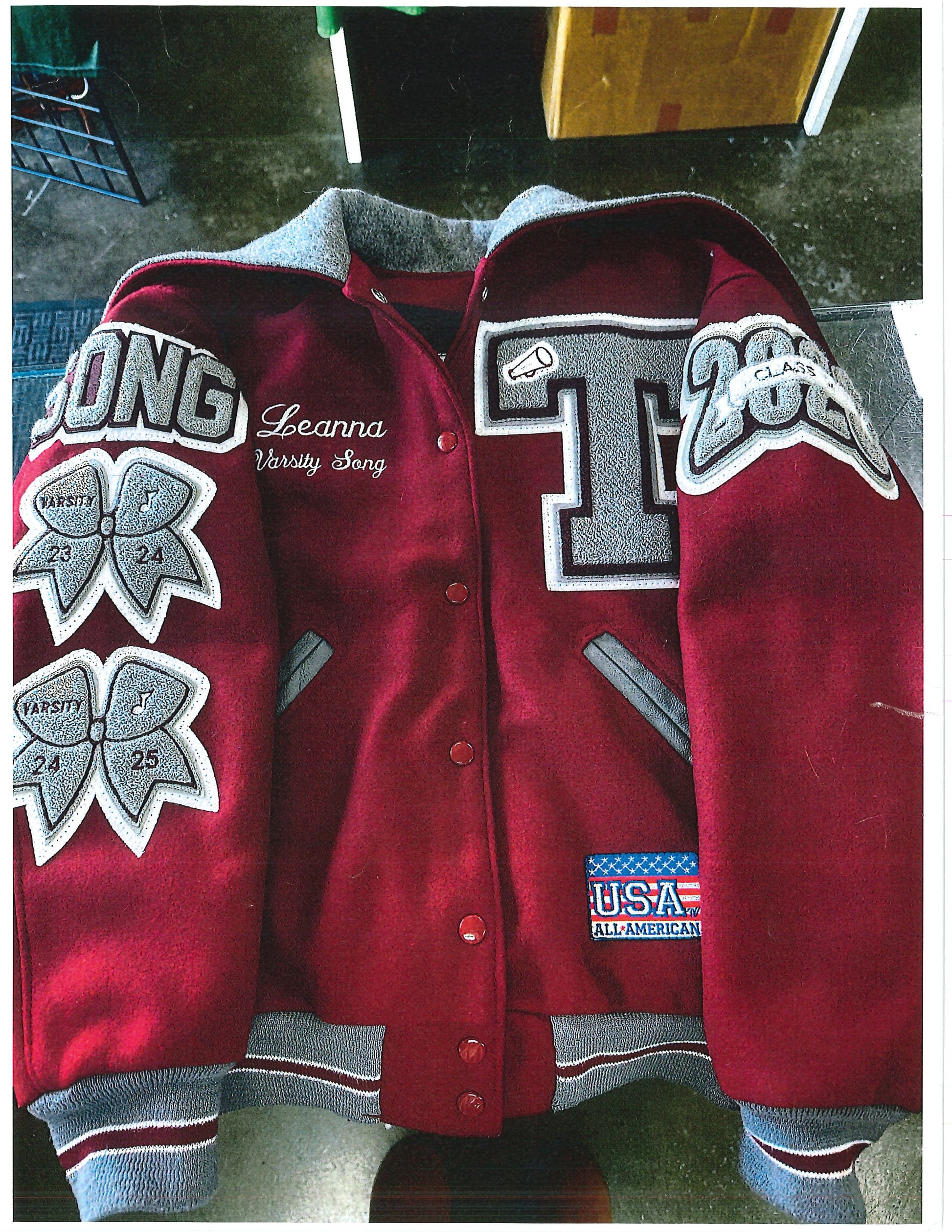A maroon varsity-style jacket with gray ribbed cuffs and collar, large gray and white embroidered letter patches, and a small USA flag patch on the chest.